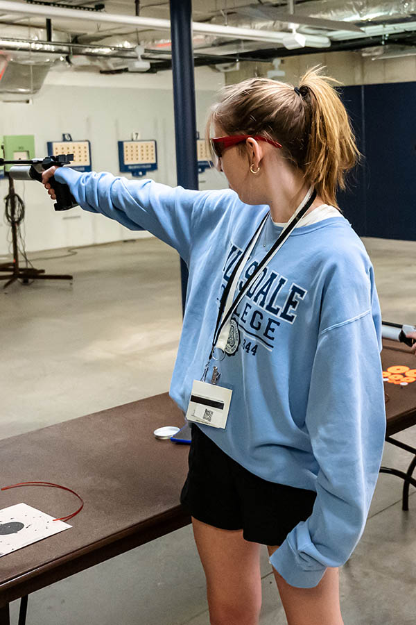 young woman in Hillsdale College light blue sweatshirt aims air rifle down range inside building