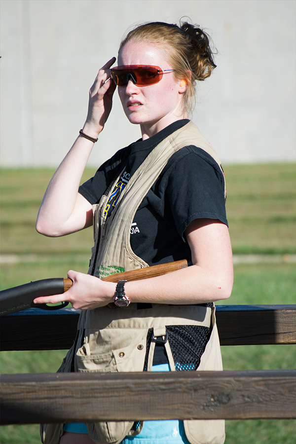 Female wearing orange safety glasses and tan vest holding shotgun.
