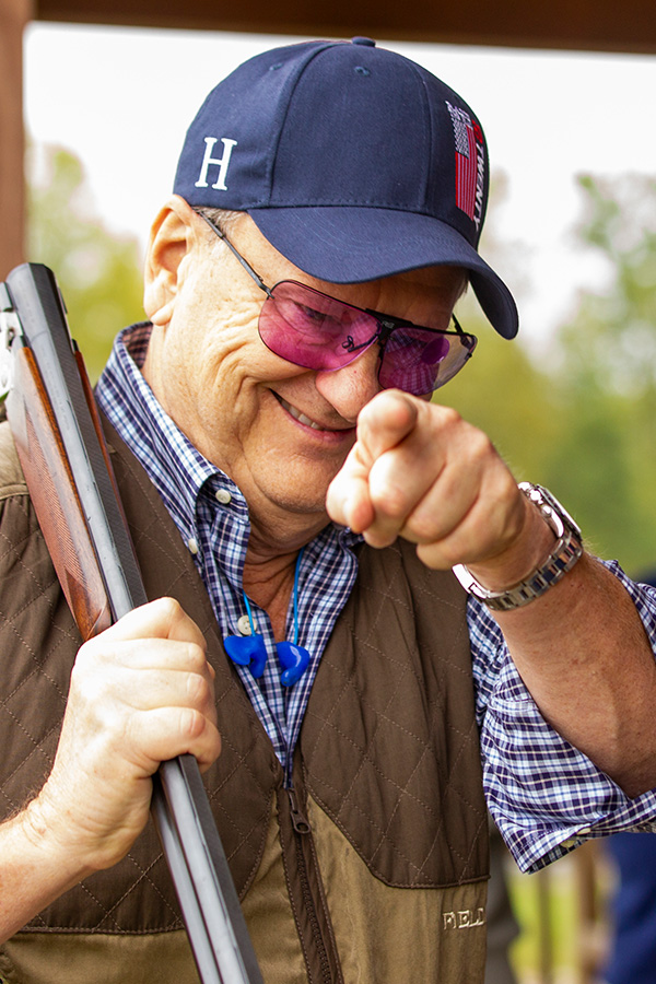 Man wearing blue ball cap holding a shotgun on his shoulder pointing and smiling at camera.