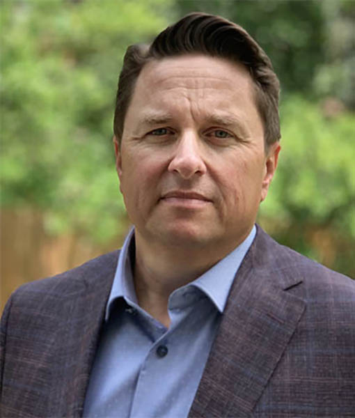 headshot of man with brown hair in plaid suit jacket and blue button down shirt looks into the camera