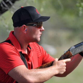 Halter shooting coach Will Hinton in a blue cap aiming hi shotgun.