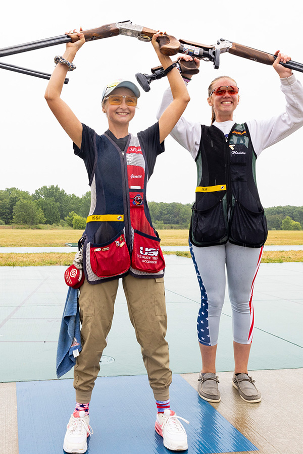 Young, female shotgun athletes proudly hold their guns above their heads.